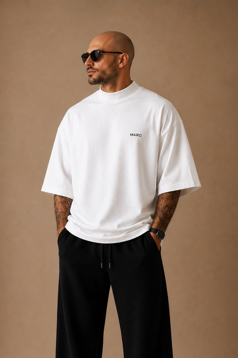 A man wearing a white short-sleeve t-shirt with a small black logo on the left chest, black sweatpants, and white sneakers, standing against a solid brown studio background.